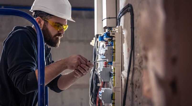 A male electrician works in a switchboard with an electrical connecting cable. A male electrician works in a switchboard with an electrical connecting cable, connects the equipment with tools.